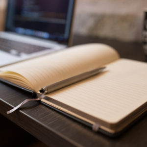 Picture of a computer, notebook, pen, and coffee mug sitting on a desk